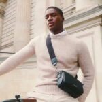 El bolso favorito de la princesa Kate está en oferta y no vas a creer los precios que tiene, hombre con jersey beige y bolso cruzado negro apoyado en una bicicleta frente a una fachada clásica