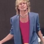 Figura con chaqueta azul y camiseta rosa en pose dramática frente a fondo oscuro, peinado rubio ochentero y accesorios llamativos; ¿Qué fue de Baby Jane? La historia detrás del clásico de terror