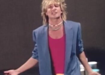 Figura con chaqueta azul y camiseta rosa en pose dramática frente a fondo oscuro, peinado rubio ochentero y accesorios llamativos; ¿Qué fue de Baby Jane? La historia detrás del clásico de terror