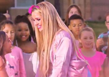Artista rubia con chaqueta rosa sonriendo y rodeada de niños en un evento al aire libre — ¿Qué fue de Aqua? Descubre la historia detrás de Barbie Girl.