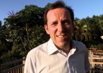 Hombre sonriente en una terraza rodeada de vegetación tropical al atardecer; El nuevo drama de la estrella de Death in Paradise arrasa con críticas increíbles y deja a los espectadores boquiabiertos.