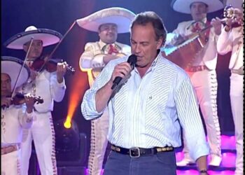 Hombre cantando en escenario acompañado por mariachis vestidos de blanco