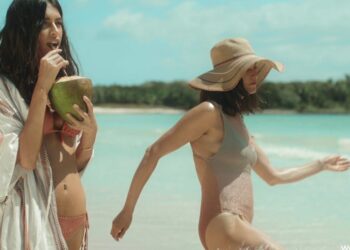 Dos mujeres jóvenes en traje de baño disfrutando en la playa, una bebiendo de un coco y otra caminando junto al mar