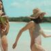 Dos mujeres jóvenes en traje de baño disfrutando en la playa, una bebiendo de un coco y otra caminando junto al mar