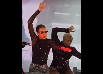 Joven artista en escenario con bailarines en trajes negros realizando una coreografía dinámica y gestos de celebración