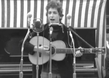 Músico joven en blanco y negro tocando la guitarra frente a varios micrófonos en un escenario al aire libre.