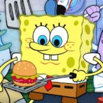 Bob Esponja sonriente sostiene una cangreburger sobre una espátula en una cocina submarina mientras otros peces observan; ¿Dónde vive Bob Esponja? Descubre la icónica ciudad de Fondo de Bikini