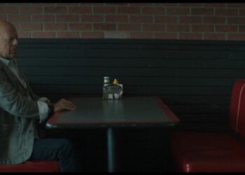 Hombre calvo sentado solo en un puesto de restaurante tipo diner frente a una mesa con condimentos.