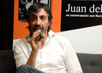 Descubre la impresionante casa de Nuria Roca y Juan del Val en Madrid: hombre con micrófono sentado en un sofá frente a un cartel promocional.