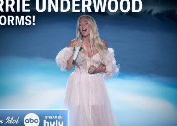 Cantante femenina en vestido blanco etéreo sosteniendo un micrófono sobre un escenario iluminado, interpretación emotiva frente a un fondo azul.