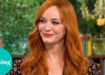 Mujer pelirroja de cabello largo y ondulado sonríe durante una entrevista en un set de televisión con fondo de plantas y estantería