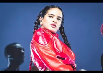 Cantante en escenario con chaqueta roja brillante y trenzas largas, mirada intensa y auriculares in-ear bajo luces azules.