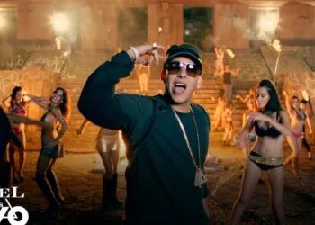 Artista urbano con gorra y gafas cantando frente a bailarinas en un escenario nocturno con iluminación cálida.