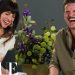 Dos personas riendo en una entrevista, una viste un vestido blanco de encaje junto a un ramo floral