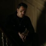 Hombre sentado en una escalera con mirada pensativa y luz tenue, ambiente íntimo y melancólico.