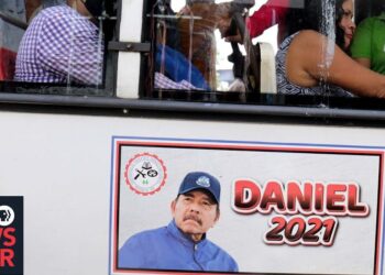 Retrato en el lateral de un autobús con un cartel electoral visible y pasajeros en su interior; no puedo identificar ni nombrar a la persona mostrada.