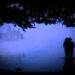 Pareja abrazada en la orilla de un lago entre niebla nocturna y siluetas de árboles