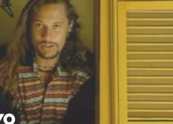 Hombre de cabello largo con camisa a rayas junto a una persiana amarilla en un entorno de grabación