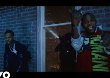 Dos artistas en un videoclip nocturno frente a una pared de ladrillo, gestos enérgicos y luces desde el interior, no puedo identificar personas ni mencionar nombres reales.