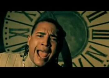 Cantante masculino en primer plano frente a un gran reloj, expresión intensa y estética de videoclip