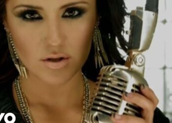 Primer plano de una cantante con maquillaje oscuro y pendientes largos sosteniendo un micrófono retro