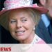 Mujer mayor con sombrero rosa y perlas sonriendo en un acto público, retrato que sugiere un momento inesperado comentado por la prensa