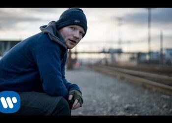 Hombre pelirrojo con gorro y chaqueta sentado junto a las vías del tren al atardecer