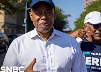 Hombre con gorra y camisa blanca muestra el pulgar arriba en una calle urbana acompañado por otra persona con gafas de sol y camiseta de campaña; no puedo identificar a la persona en la imagen.