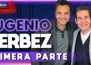 Dos hombres sonrientes en un set de entrevista con letreros luminosos y un cartel promocional que anuncia la primera parte.