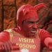 Hombre disfrazado de diablo en traje rojo sosteniendo un cartel que dice 'VISITA KOSOVO' durante un sketch televisivo