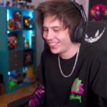 Persona joven sonriendo con auriculares en un estudio de streaming, silla gamer y estantería iluminada.