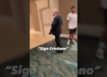 Hombre mayor con traje caminando por un pasillo mientras otro joven lo observa, con texto superpuesto "Sign Cristiano".
