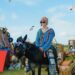 Persona joven con suéter colorido montada en un burro adornado con gafas de sol y notas musicales en un ambiente festival al aire libre; Hijos de Kratos: descubre la historia y legado de Atreus y Calíope