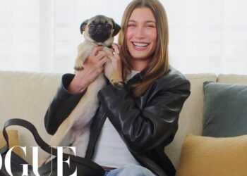 Mujer sonriente sentada en un sofá sosteniendo un perro pequeño, look casual con chaqueta de cuero y ambiente hogareño.