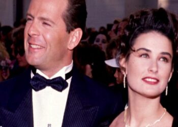 Pareja elegante en alfombra roja de gala, mujer con vestido y joyas junto a hombre con esmoquin, mirada esperanzada.