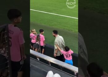 Niños con camisetas rosas y números en la grada de un estadio observando el campo