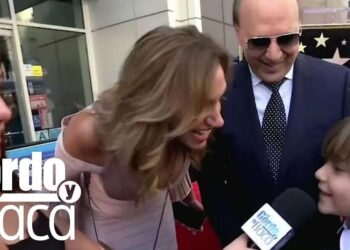 Madre sonriente inclinándose para hablar con un niño en la alfombra roja durante una entrevista con micrófono visible