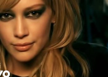 Primer plano de mujer rubia con maquillaje ahumado, pendientes y mirada intensa sobre fondo oscuro