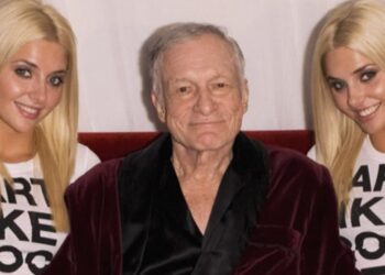 Hombre mayor sentado en bata entre dos mujeres rubias sonrientes en un ambiente de celebración