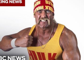 Hombre musculoso con pañuelo rojo y camiseta amarilla posando con gesto desafiante junto a rótulos de 'Breaking News' y logo de NBC News