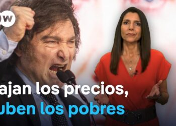 Orador enfático en un podio junto a presentadora y el titular en pantalla 'Bajan los precios, suben los pobres'.