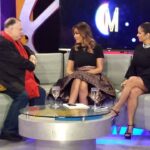 Invitado y presentadoras sentados en un set de televisión moderno con sofá y mesa de cristal, ¿Qué fue de César Vidal? Descubre su vida y actualidad impactante