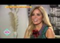 Mujer rubia sonriente en una entrevista televisiva, con vestido verde y broche floral, ambiente cálido de estudio.