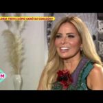 Mujer rubia sonriente en una entrevista televisiva, con vestido verde y broche floral, ambiente cálido de estudio.