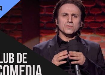 Presentador en traje oscuro con micrófono en un escenario de club de comedia frente a un fondo de ladrillo