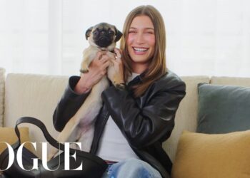 Mujer sonriente sostiene un perro pug en el sofá, respondiendo con humor a rumores y acusaciones en redes sociales.