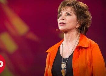 Mujer adulta hablando en un escenario con chaqueta naranja y micrófono de diadema, ¿Dónde vive Isabel Allende? Descubre su hogar y estilo de vida