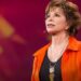 Mujer adulta hablando en un escenario con chaqueta naranja y micrófono de diadema, ¿Dónde vive Isabel Allende? Descubre su hogar y estilo de vida