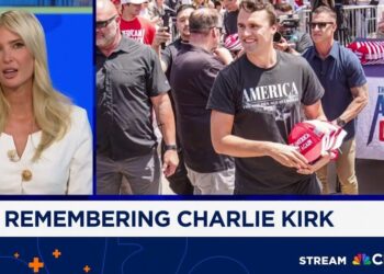 Mujer rubia con chaqueta blanca en pantalla de noticias junto a hombre sonriente con camiseta 'AMERICA' entregando gorras rojas en un mitin