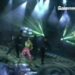 Artista en escenario con falda rosa y dos bailarines bajo intensas luces verdes y efectos de estudio, ambiente de show televisivo